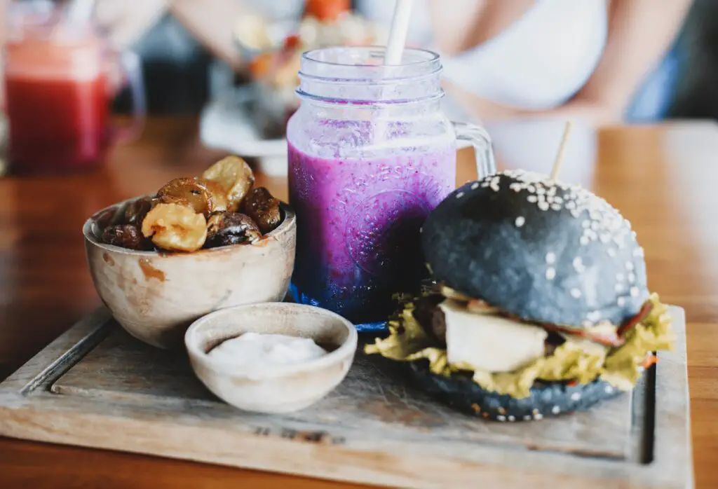 Delicious black burger with freshly made purple smoothie and wooden bowl of dried bananas and dates served on wooden board in cafe